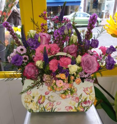 A vibrant bouquet with pink, purple, and yellow flowers in a decorative floral bag.