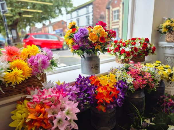 Colourful artificial flowers in pots displayed in a shop window with a street view outside.