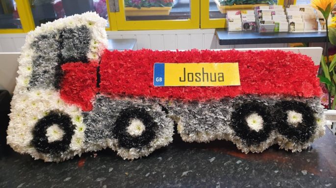 A floral arrangement shaped like a red and grey truck with a yellow sign saying "Joshua."
