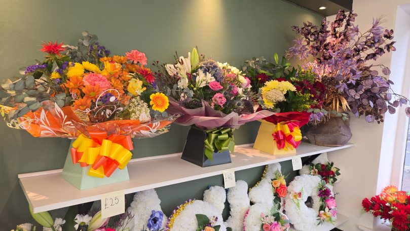 Brightly coloured flower arrangements displayed on a shelf against a green wall.