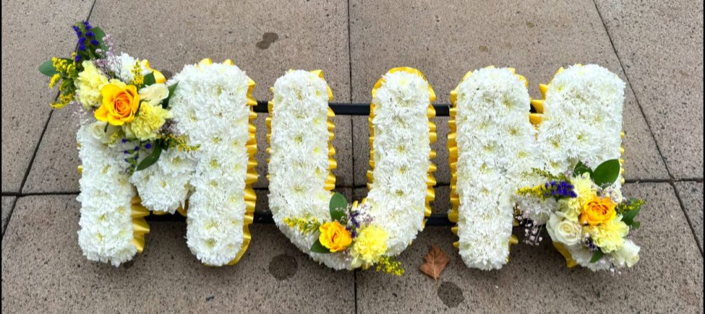 Floral tribute spelling "MUM" with yellow and purple flowers.