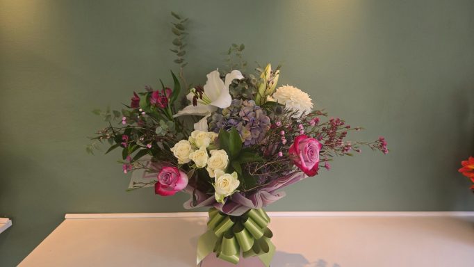 Colourful floral arrangement with pink roses, lilies, and greenery against a green background.